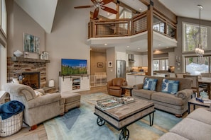 A spacious living area with a fireplace, comfortable seating, a large TV, and a kitchen in the background.