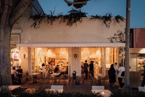 An outdoor dining area with tables and chairs, a building entrance, and a tree.