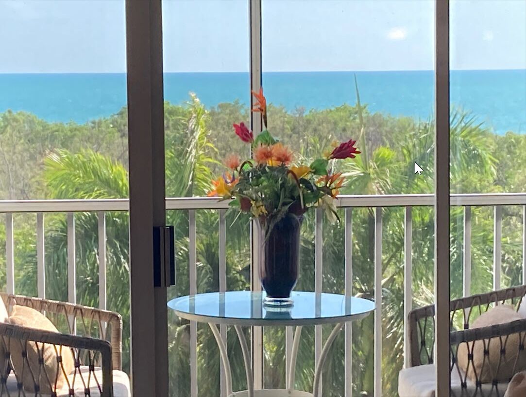 Un balcón con una mesa y sillas de vidrio, un jarrón con flores y vista al océano.