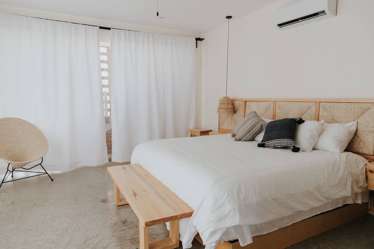 A bedroom with a wooden bed, white bedding, a wooden bench, a wicker chair, and a small bedside table.