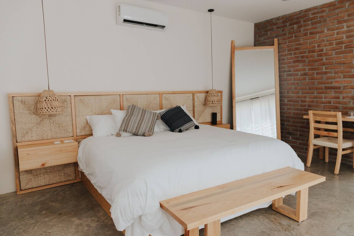 A bedroom with a wooden bed, a bench, a wooden chair, a white air conditioner, and a brick wall.