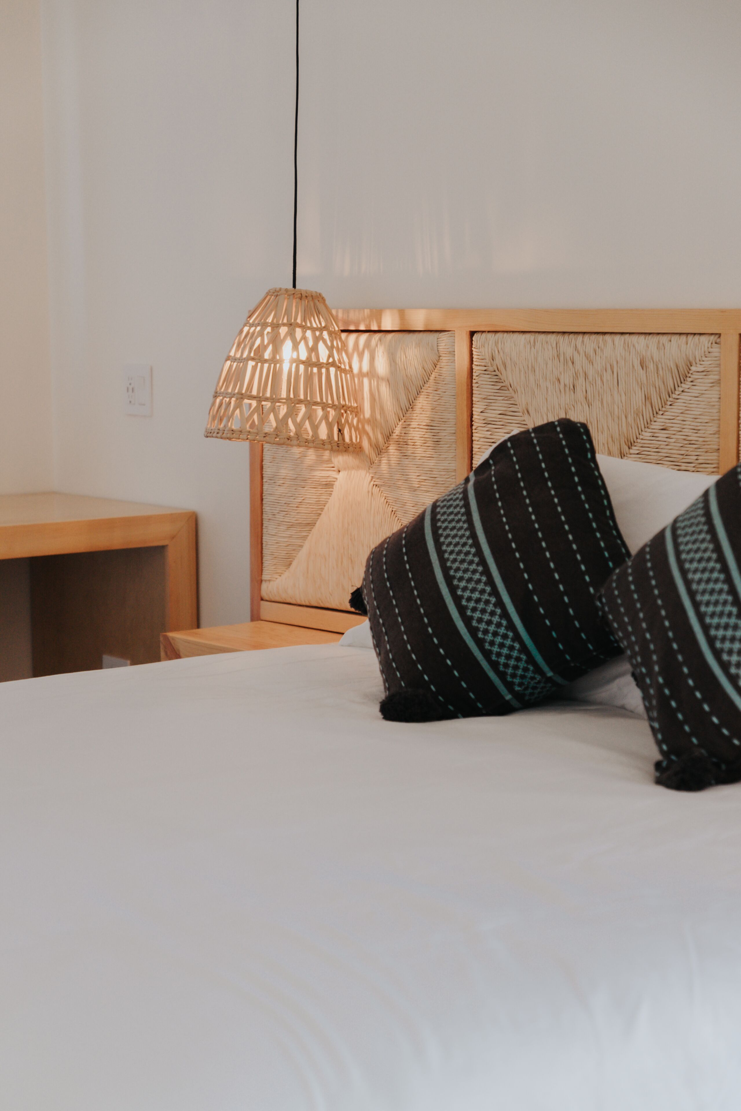 Una cama bien hecha con almohadas a rayas, un cabecero tejido y una lámpara colgante.