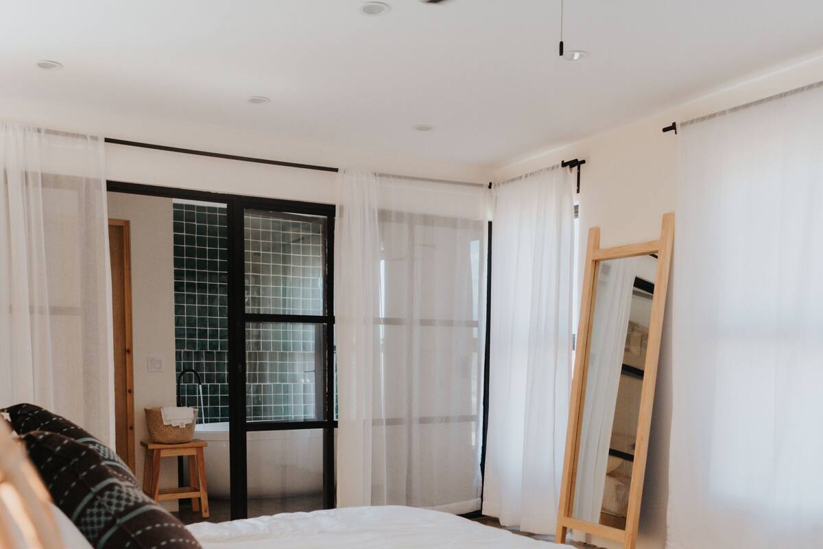 A bedroom with a bed, a ceiling fan, a window with curtains, and a mirror leaning against the wall.