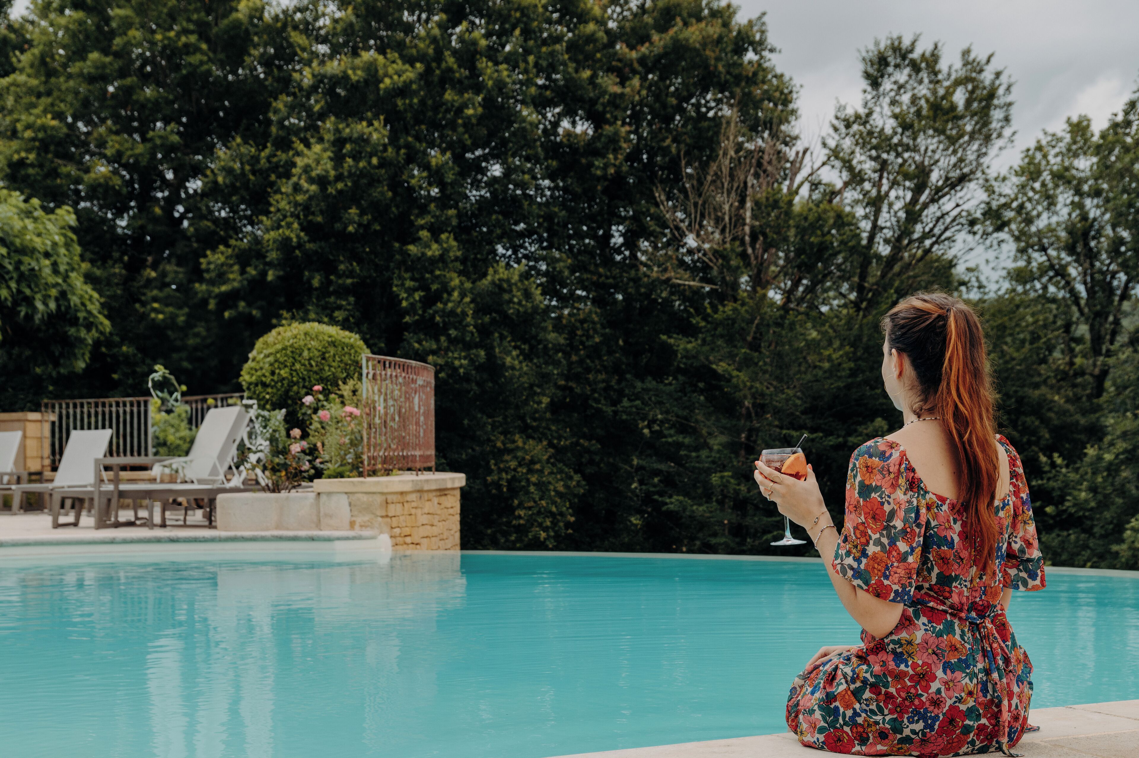 A woman sitting by a pool with a drink, surrounded by greenery and a wooden deck with lounge chairs.