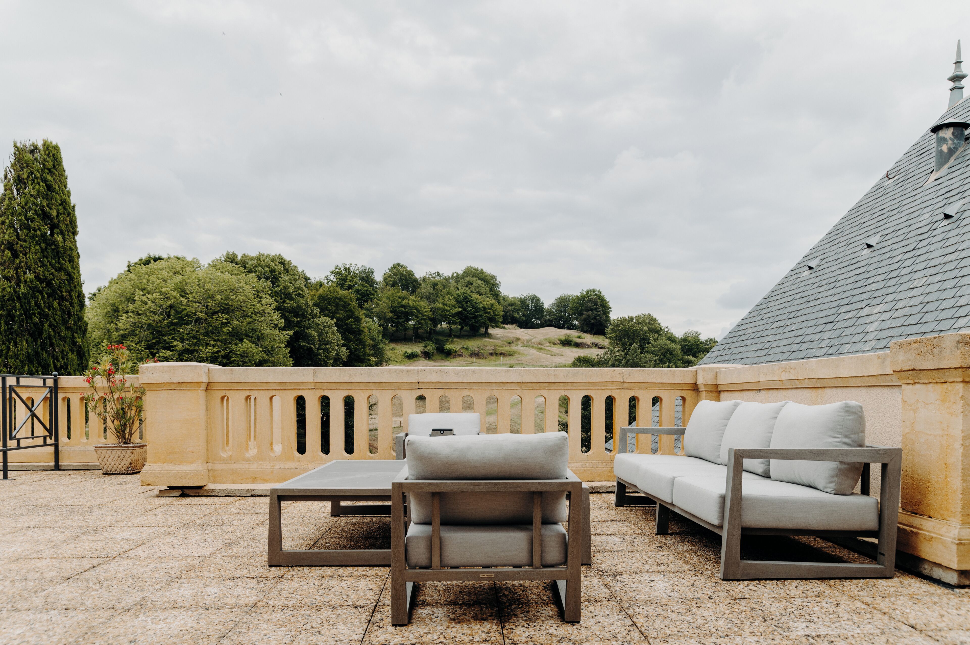 Una terraza con mobiliario exterior, incluyendo un sofá y sillas, con vista a un jardín ajardinado con árboles y una colina.