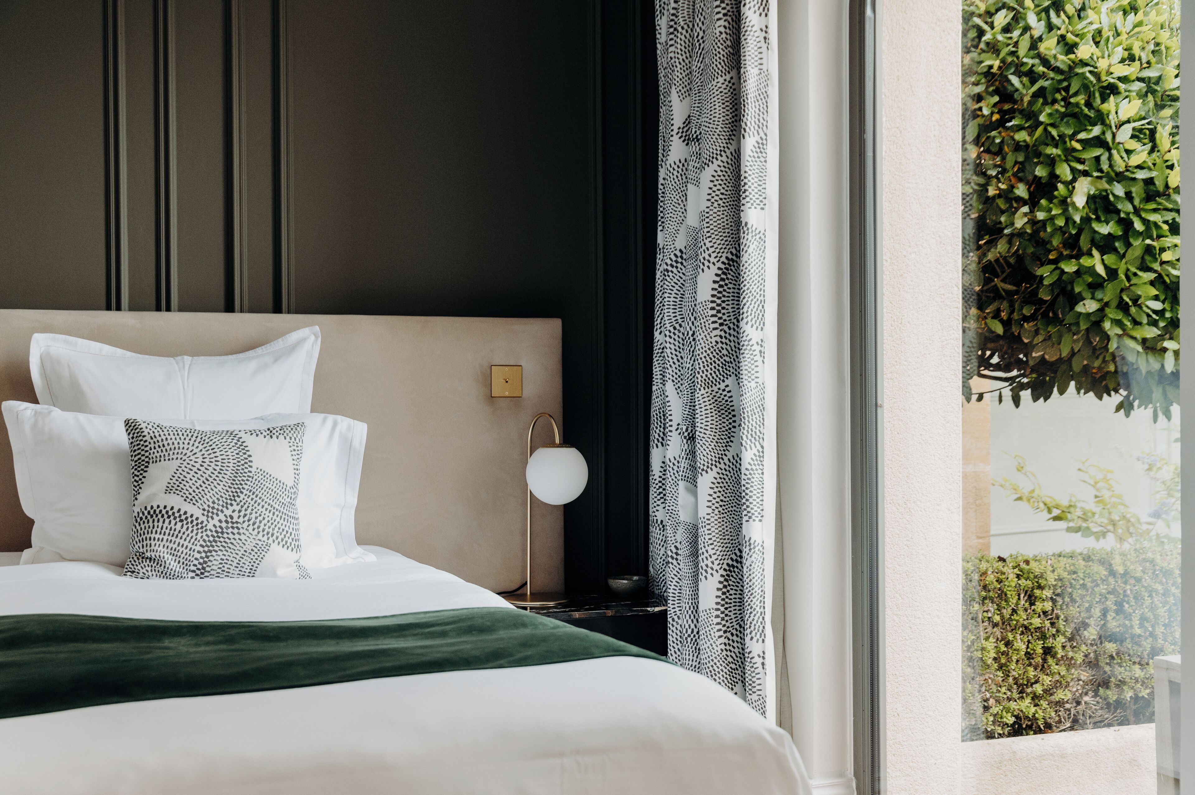 Una cama bien hecha con ropa blanca y un plaid verde, una mesita de noche con una lámpara, y una ventana con vistas a la vegetación.