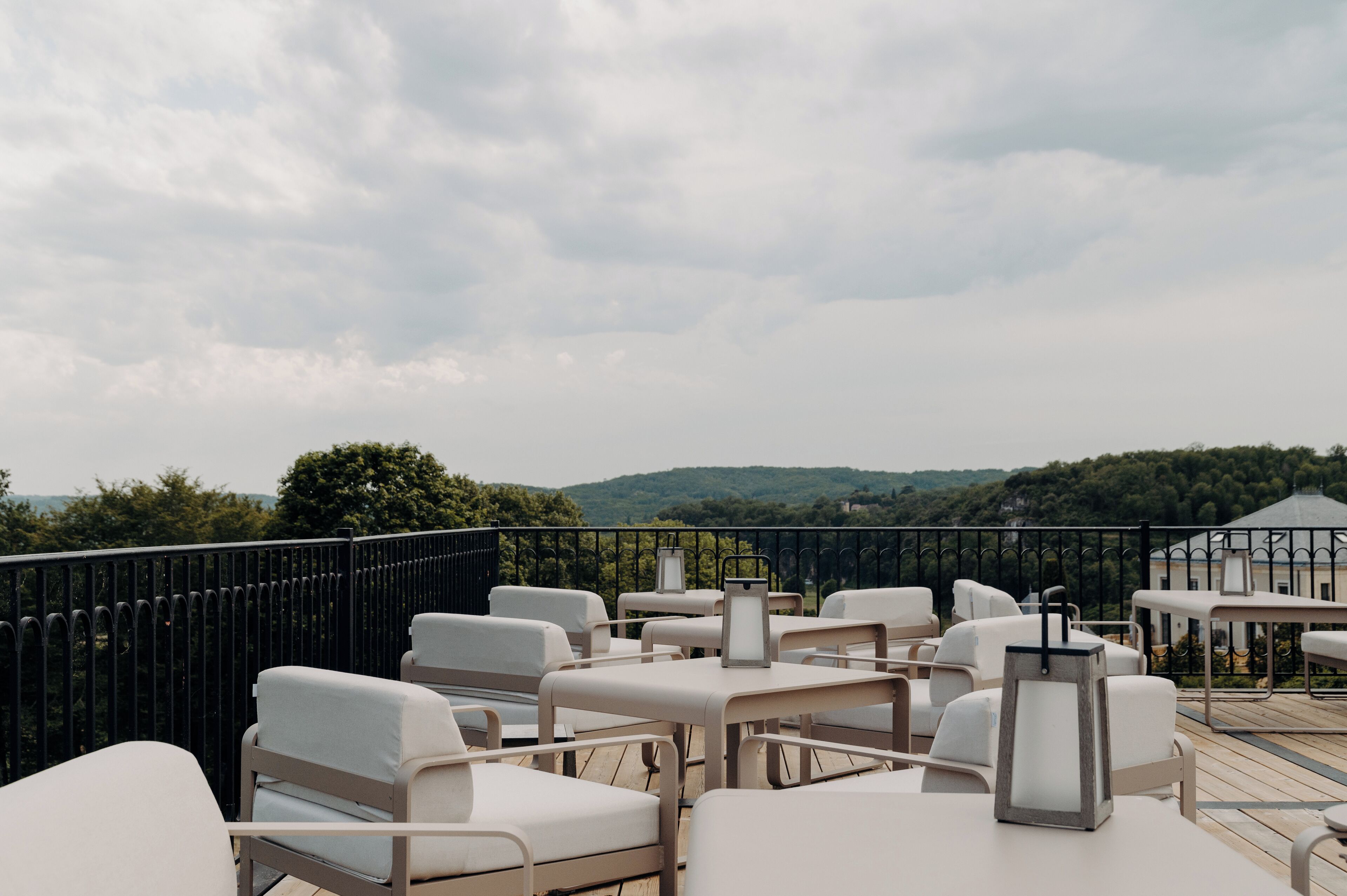 An outdoor seating area with white cushioned chairs and tables, a metal railing, and a view of green hills in the background.