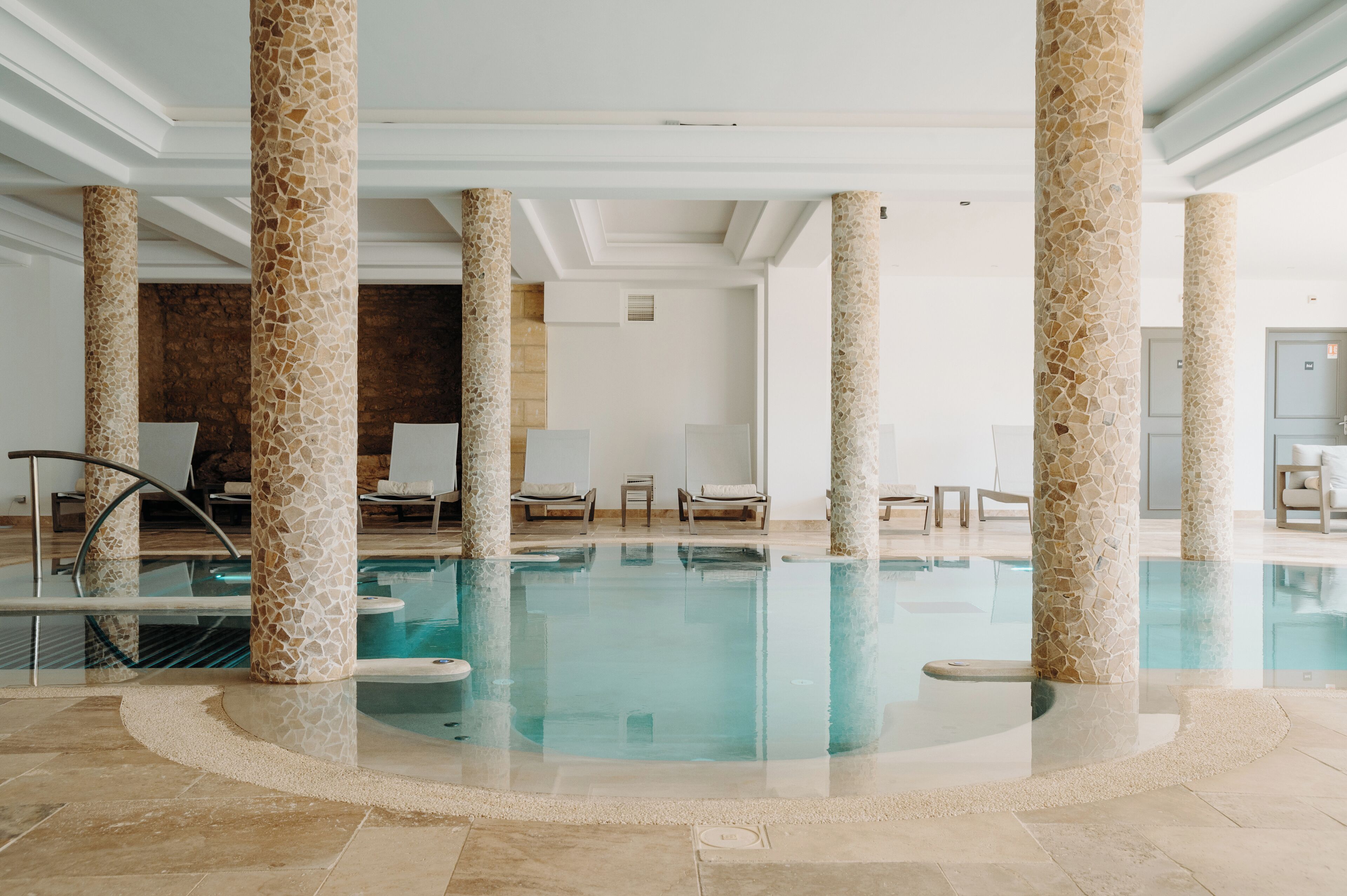 An indoor swimming pool with mosaic columns, lounge chairs, and a stone wall in the background.