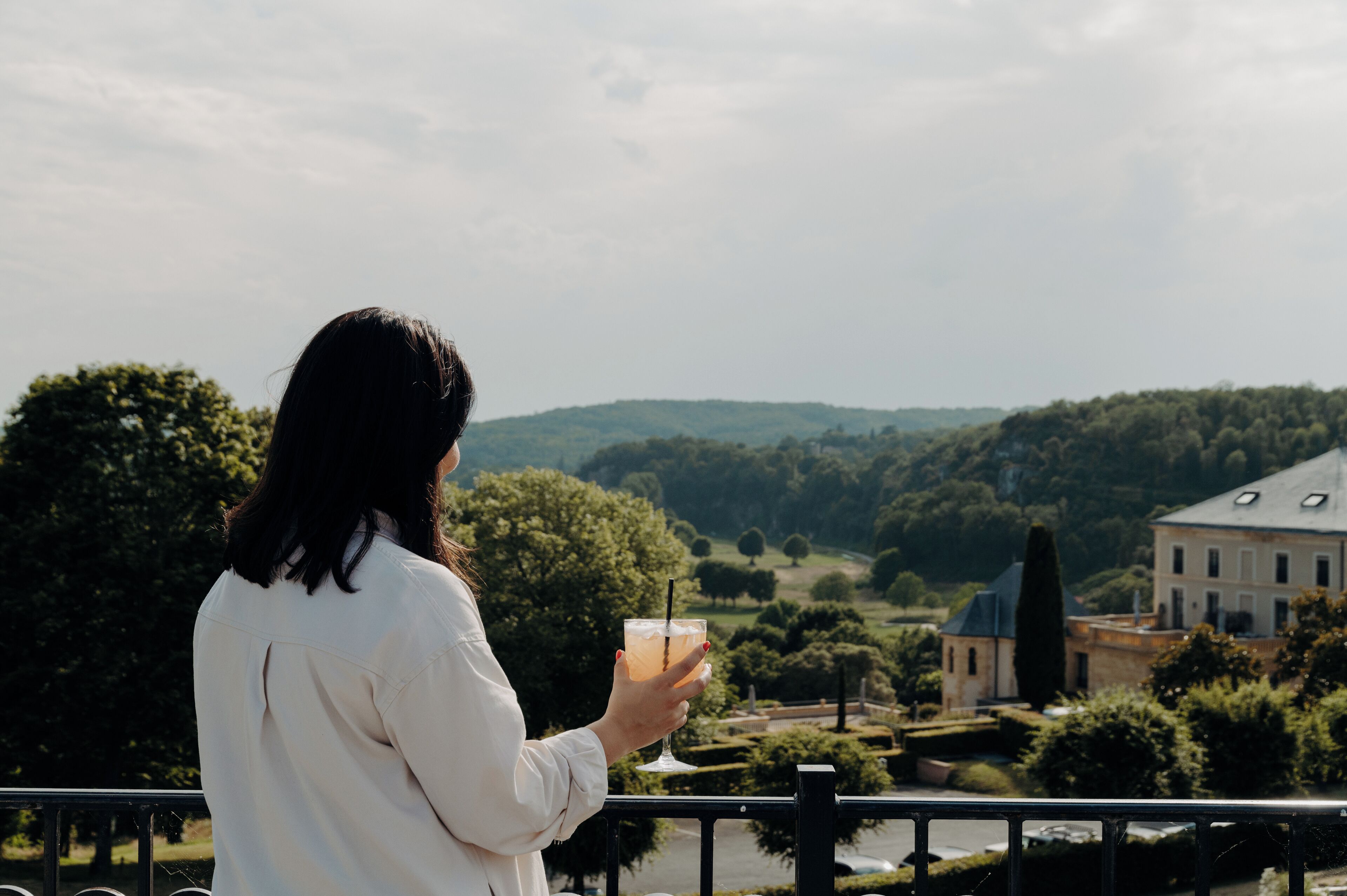 Una persona sosteniendo una bebida con vistas a un paisaje pintoresco, con un edificio a lo lejos.