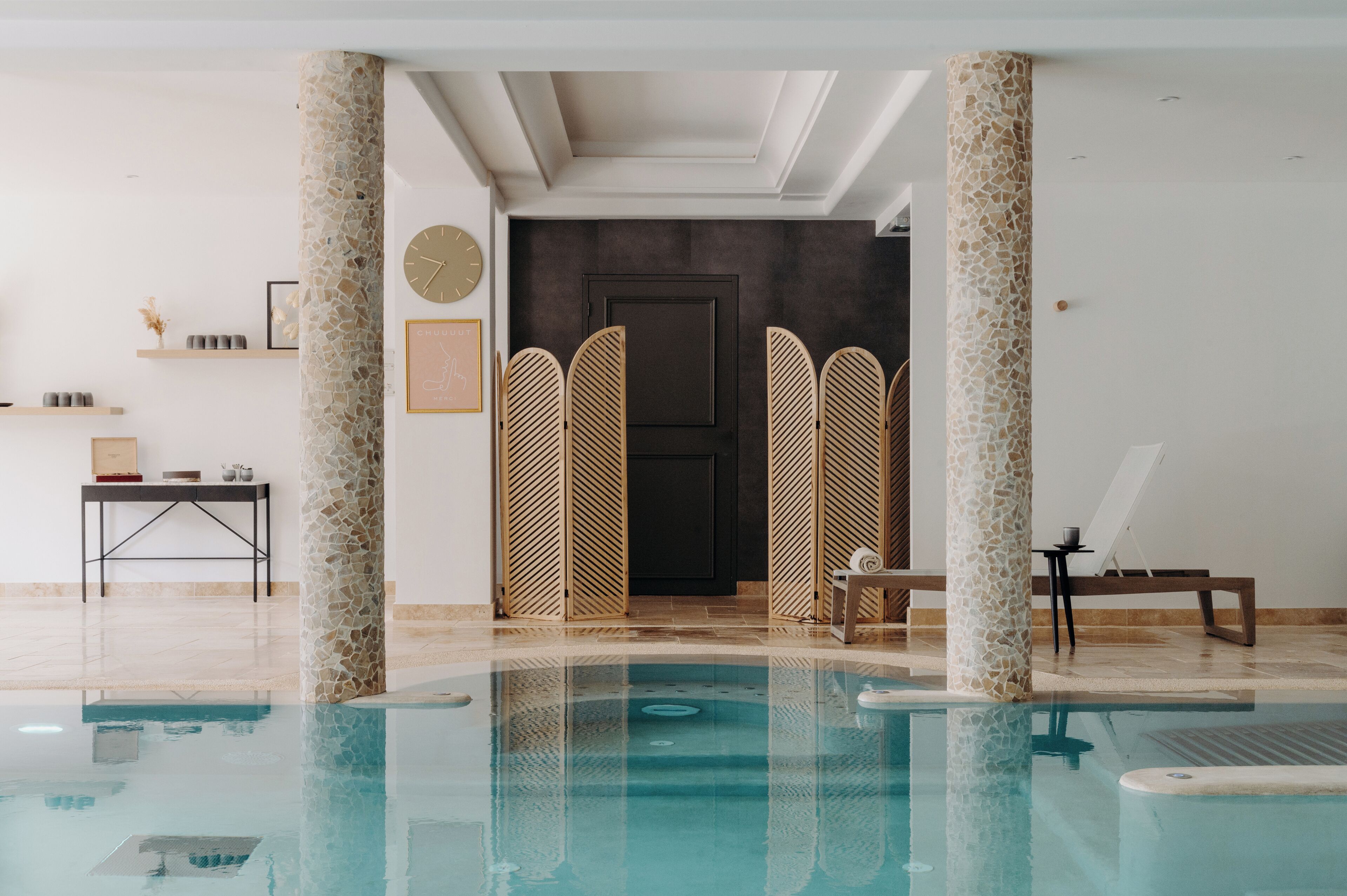 Un espace piscine intérieur moderne, doté d’une piscine à eau claire, de colonnes en pierre, d’un comptoir d’accueil et d’une porte donnant sur une autre pièce.