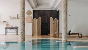 Un espace piscine intérieur moderne, doté d’une piscine à eau claire, de colonnes en pierre, d’un comptoir d’accueil et d’une porte donnant sur une autre pièce.