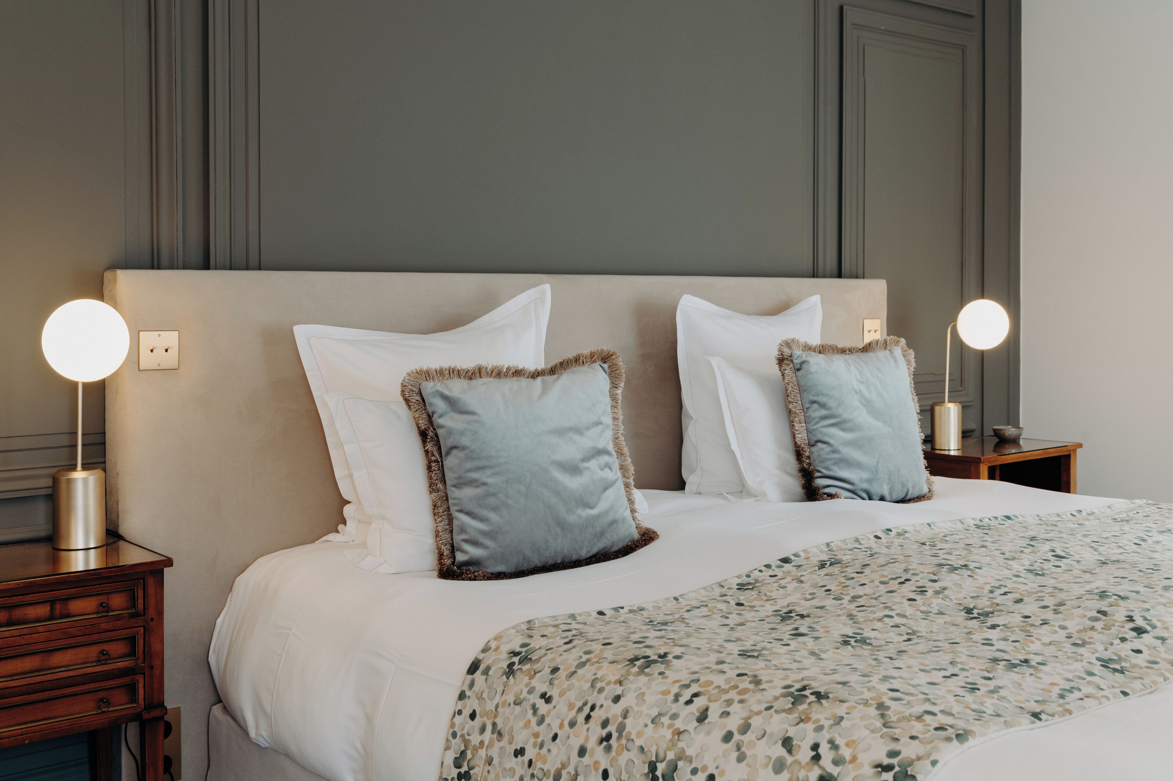 A neatly made bed with white bedding and two decorative pillows, flanked by bedside tables with round lamps.