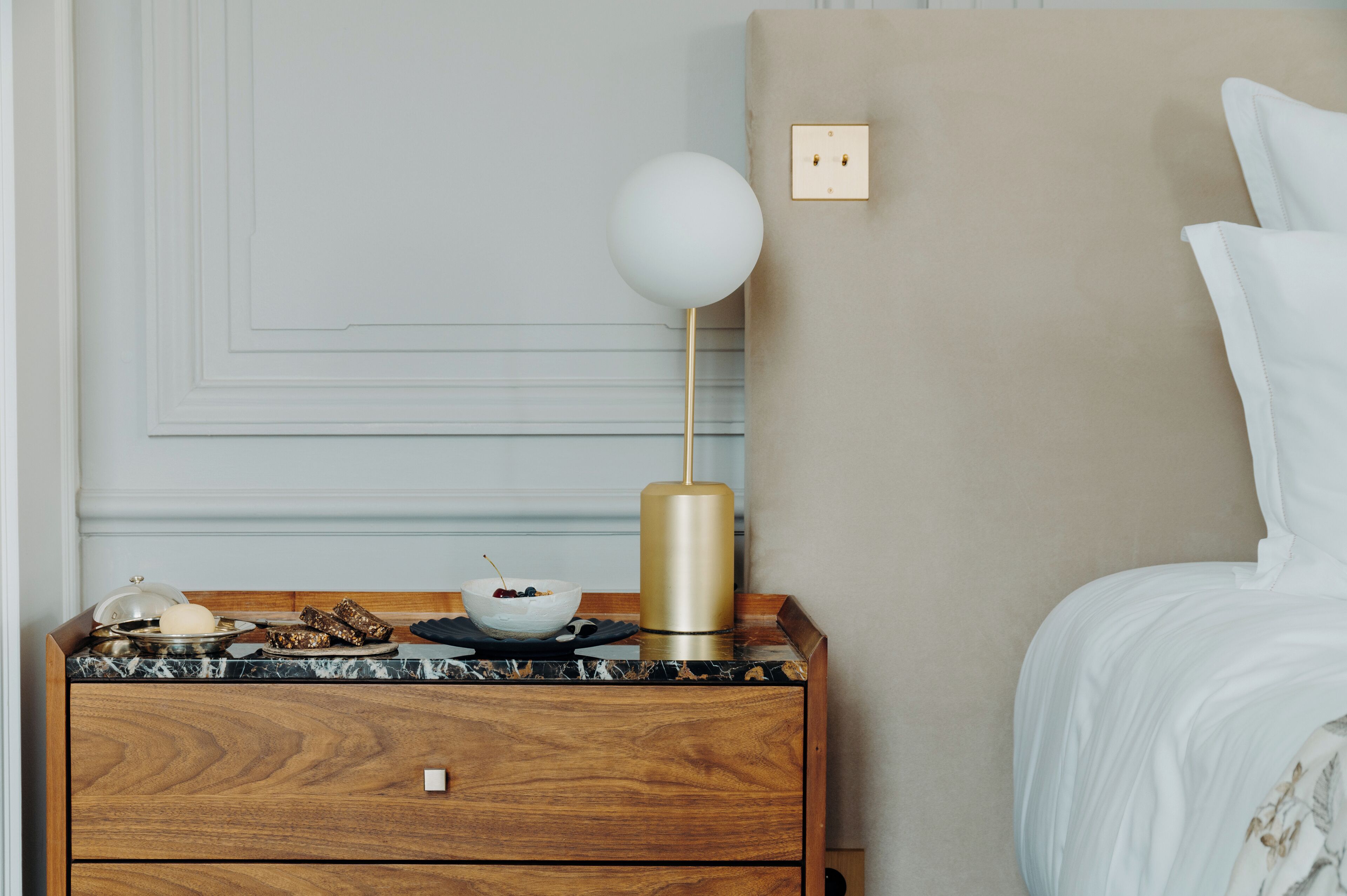 Un dormitorio ordenado con una mesita de noche de madera, un frutero, una lámpara y una cama con ropa blanca.