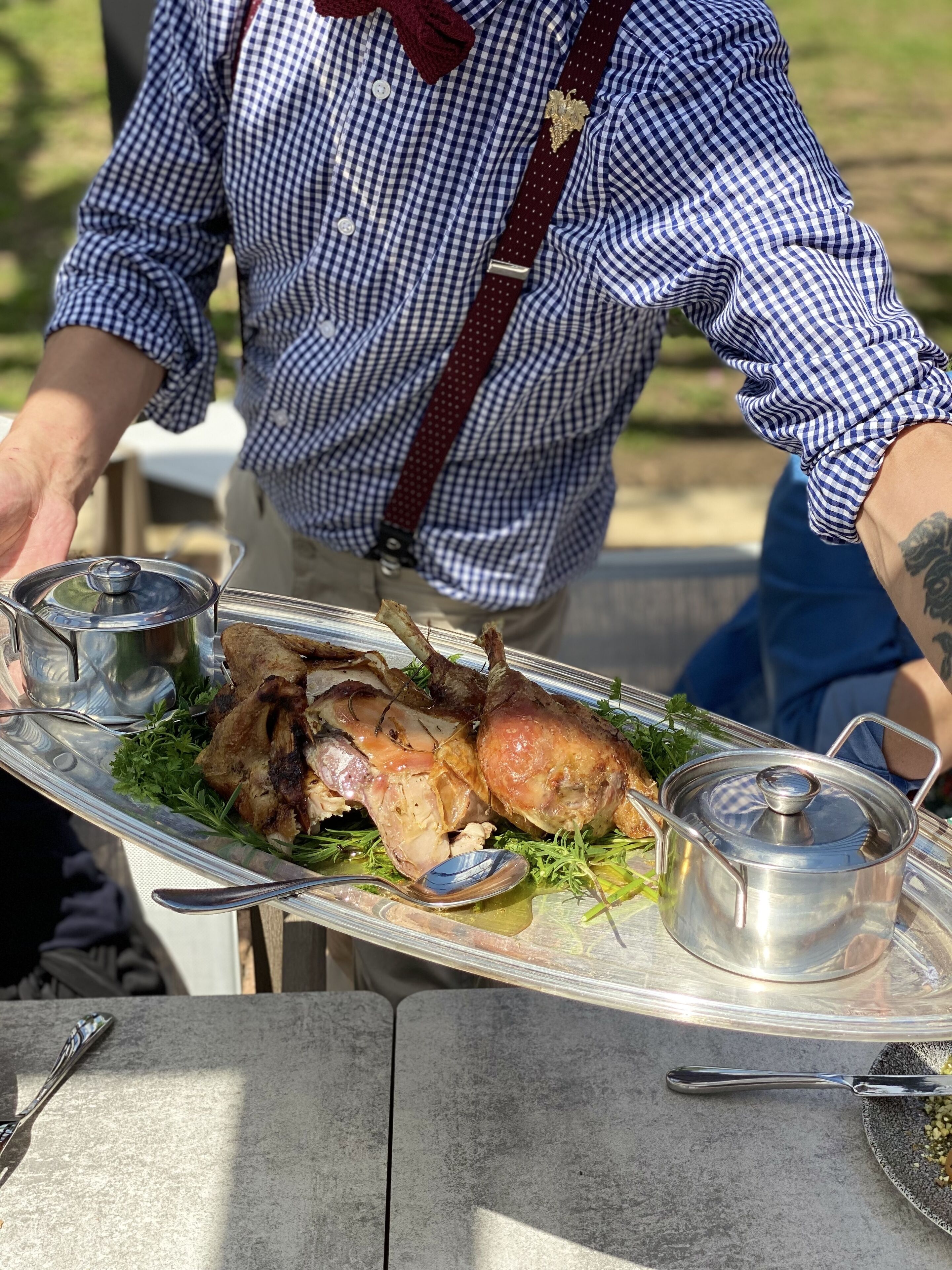 Une personne servant un poulet rôti sur un plat en argent, muni d’un couvercle et accompagné d’une cuillère, à l’extérieur.