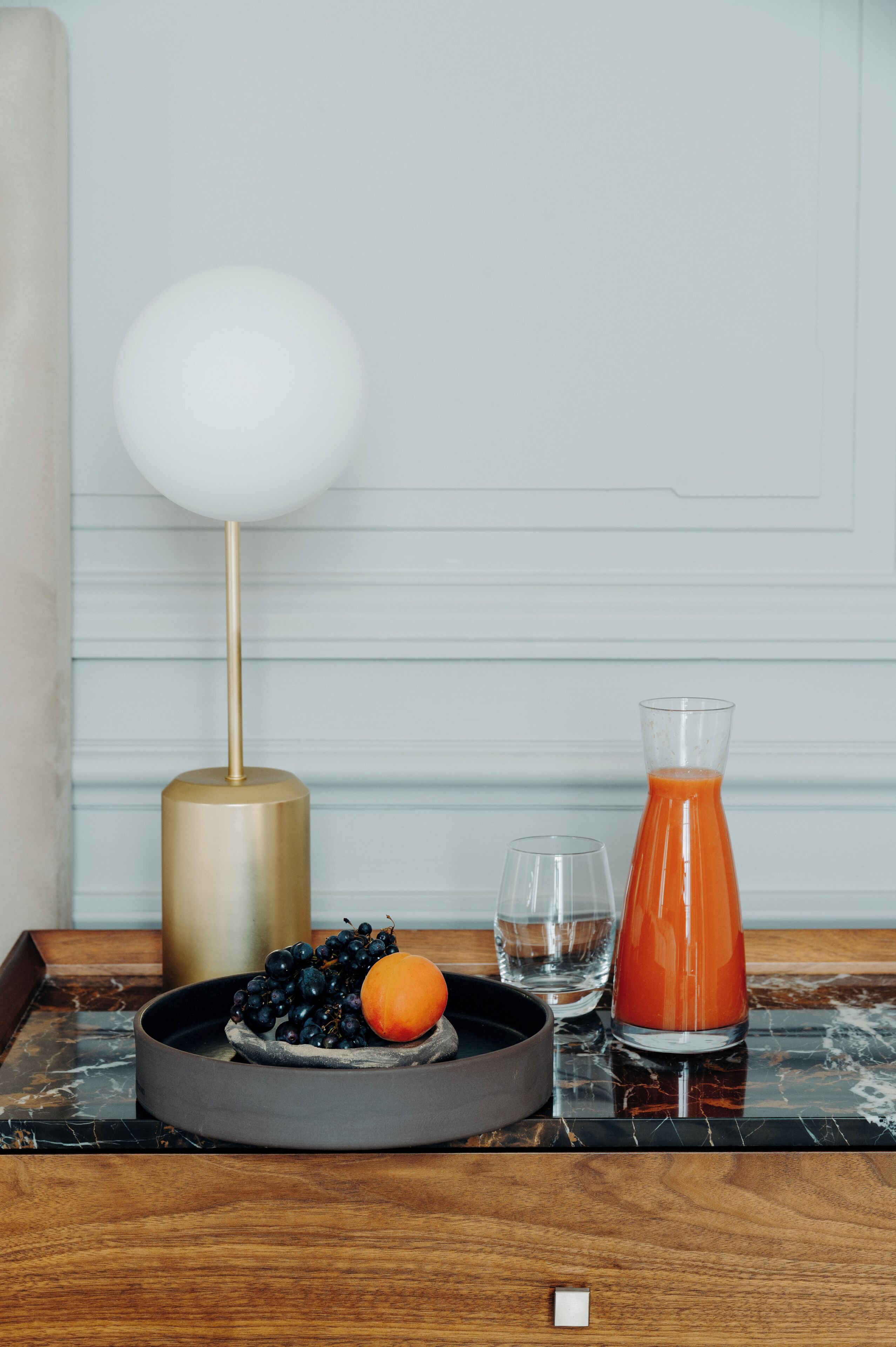 Una mesa de comedor con un frutero que contiene uvas y una naranja, un vaso de agua y un vaso de jugo de naranja.