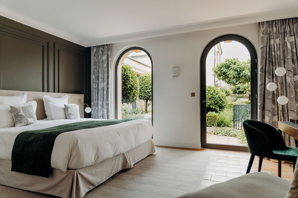 Une chambre avec un grand lit, une vue sur un jardin accessible par un passage en arc de cercle, et un coin repas avec une chaise et une table.