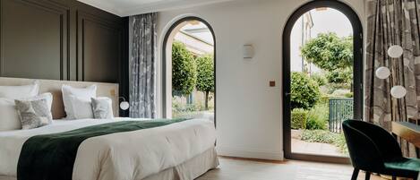 A bedroom with a large bed, a view through an arched doorway to a garden, and a dining area with a chair and table.