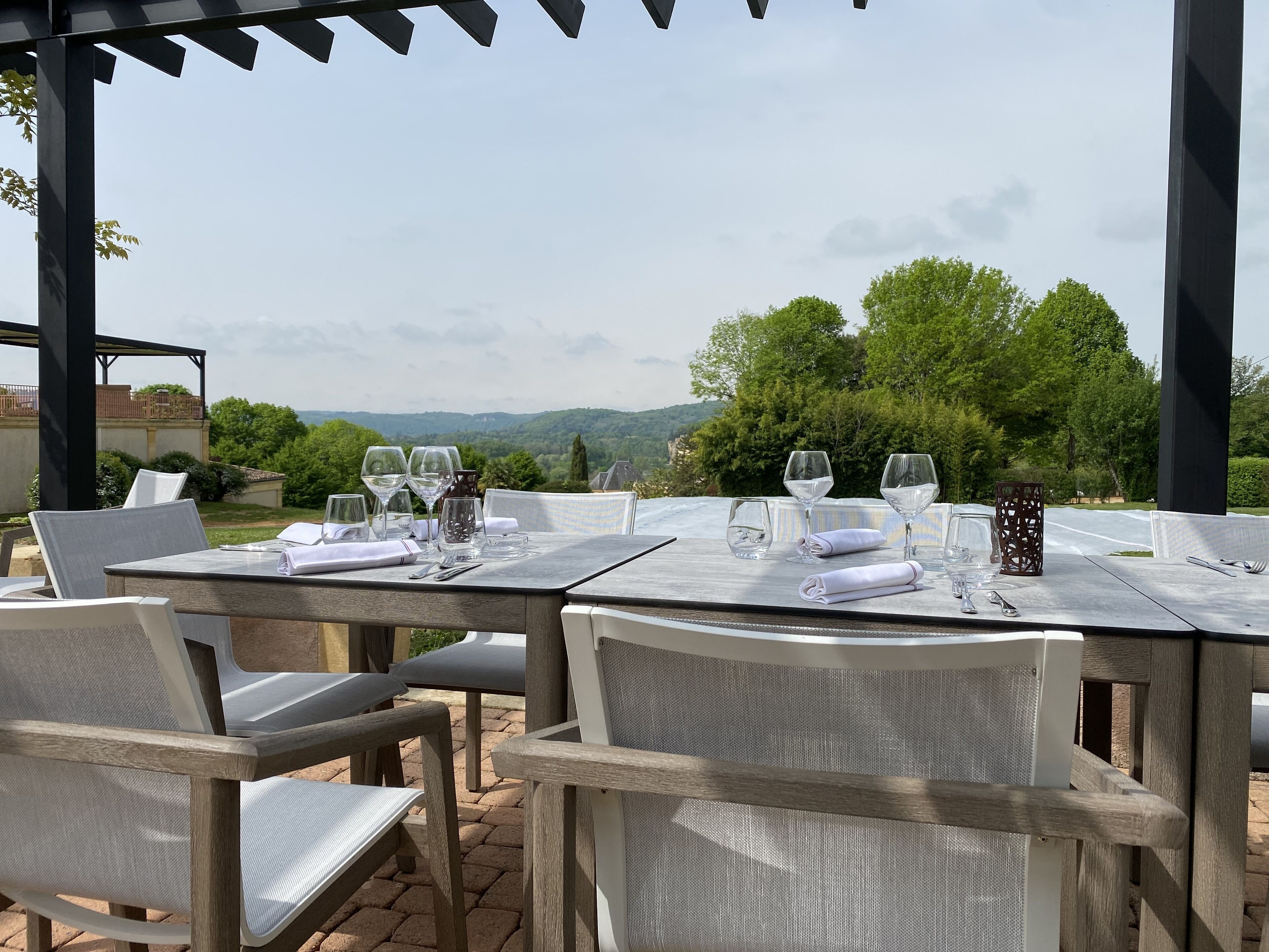 An outdoor dining area with tables set for two, equipped with wine glasses and cutlery, overlooking a scenic landscape.