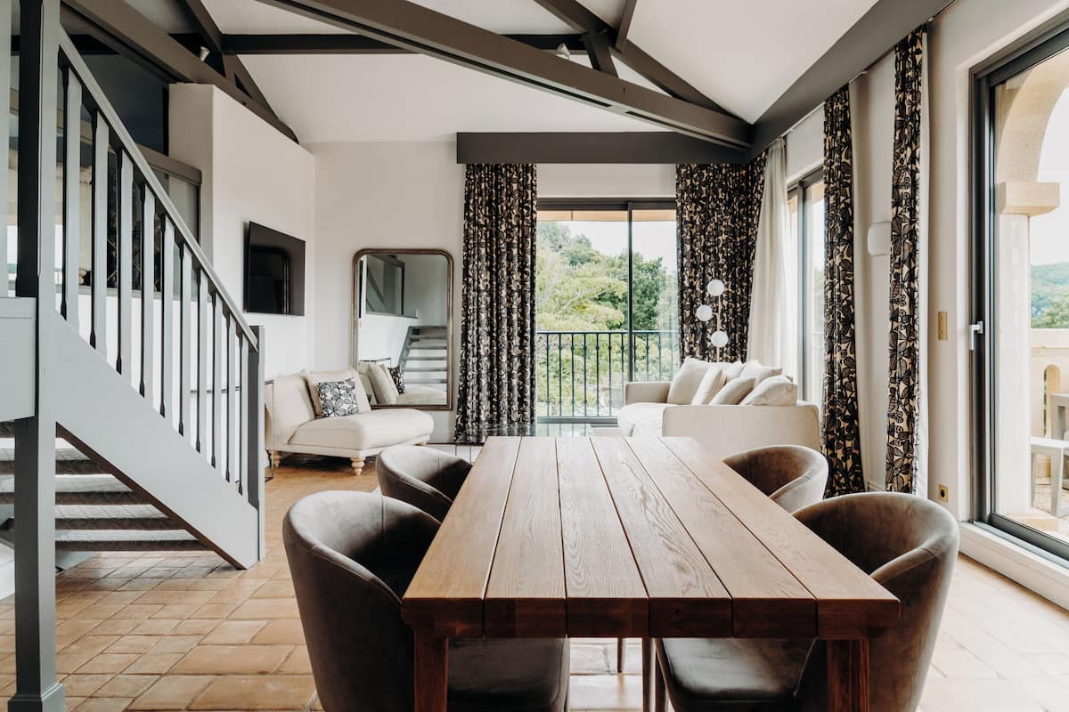 Un vaste espace de vie agrémenté d’une table en bois et de sièges confortables, d’un escalier et de grandes fenêtres dotées de rideaux.