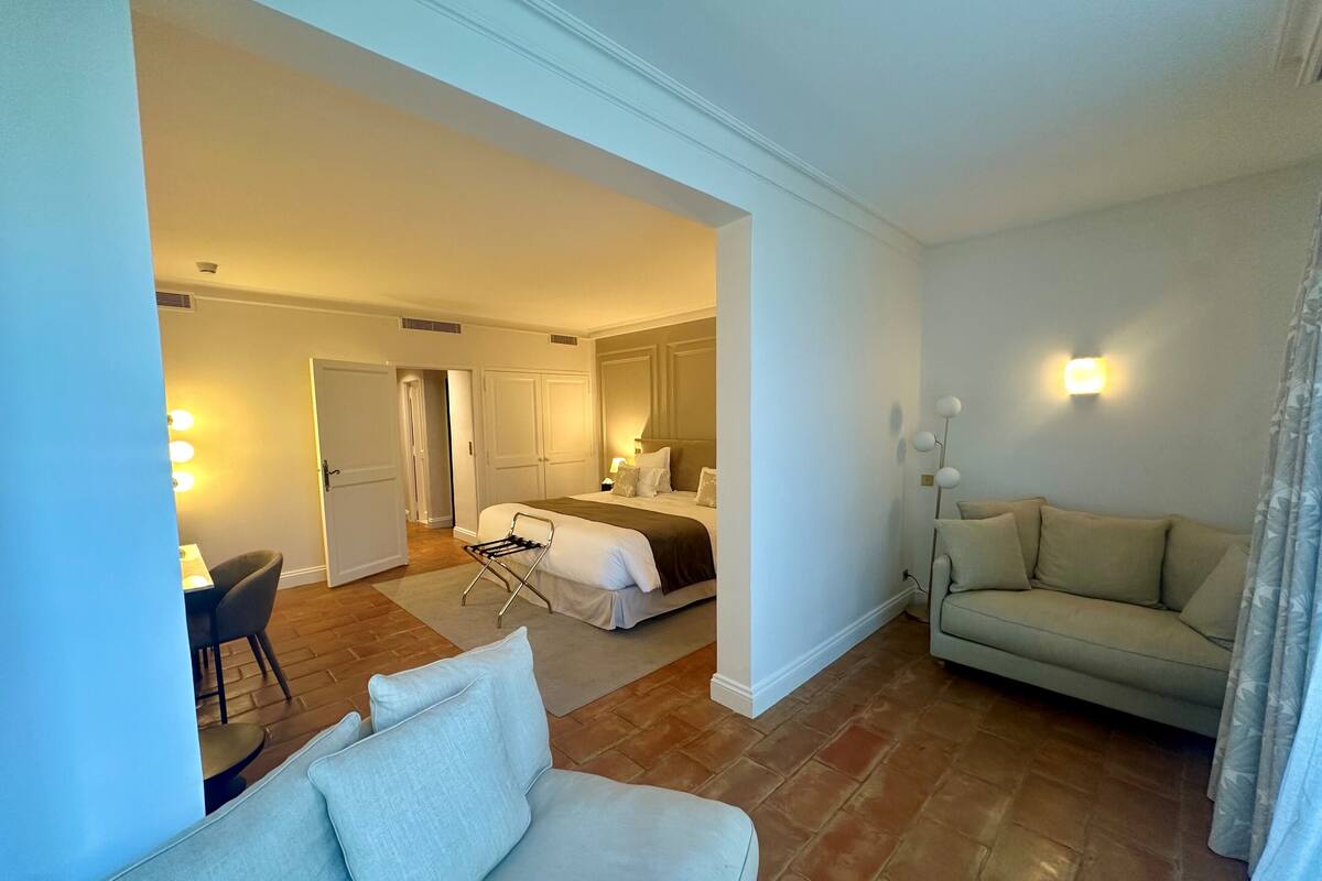 Une chambre d’hôtel comprenant un lit, un coin salon avec un canapé, une petite table et une chaise. Une porte donne accès à une autre pièce.