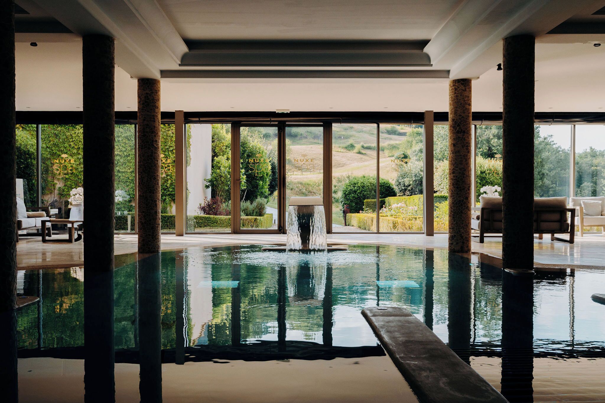 Una piscina interior con paredes de vidrio transparente, un elemento acuático en el centro y vistas a un jardín ajardinado.