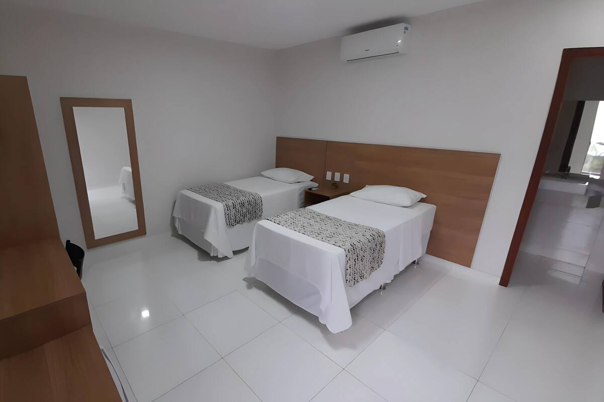 A hotel room with two single beds, a wooden headboard, a flat-screen TV, a mirror, and a white air conditioning unit.