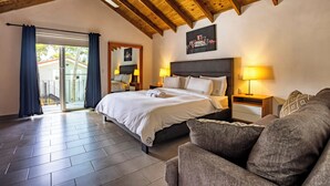 Un dormitorio con una cama grande, dos mesitas de noche con lámparas, un sofá gris, vigas de madera en el techo y vistas a través de un ventanal.