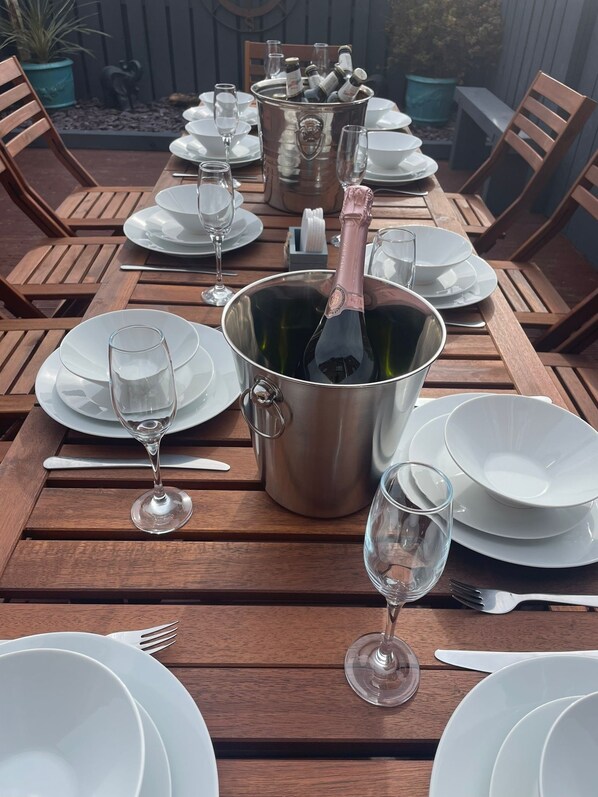 A wooden dining table set with white plates, glasses, and cutlery, a champagne bottle in a bucket, and a potted plant in the background. - The Glynn, Portrush. Deluxe holiday home, 8 beds 12 guests 5 bedrooms. (Portrush)