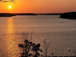 Un coucher de soleil sur une étendue d’eau calme, avec les silhouettes d’arbres et une côte lointaine.
