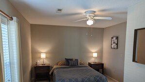 Une chambre à coucher avec un lit, deux tables de chevet avec des lampes, un ventilateur de plafond et une fenêtre avec des stores.