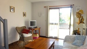 Uma sala de estar com mesa de centro de madeira, sofá de vime e uma televisão sobre uma mesinha lateral.