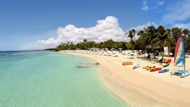 A beach with clear turquoise water, a sandy shore, and several colorful kayaks and a sailboat.
