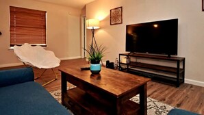 A living room with a wooden coffee table, blue sofas, a white chair, a TV on a wooden stand, and a plant on the table. - Cozy Spacious Corporate Housing 4 BR|2.5 Bath|Loft|Office|Near Tesla (Austin)