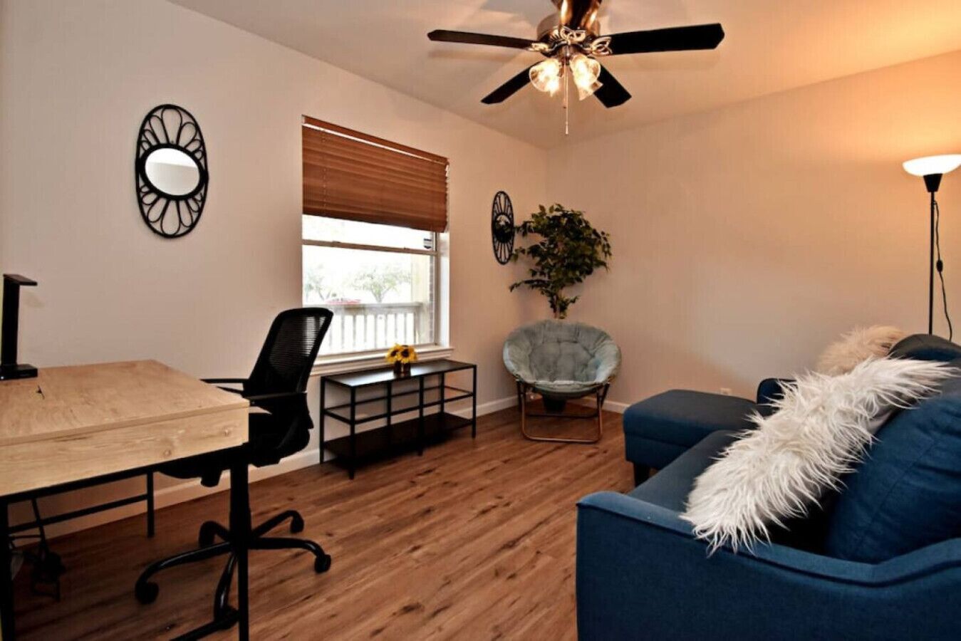 A modern living room with a dining table, a blue sofa, a chair, a ceiling fan, and a window with blinds.
