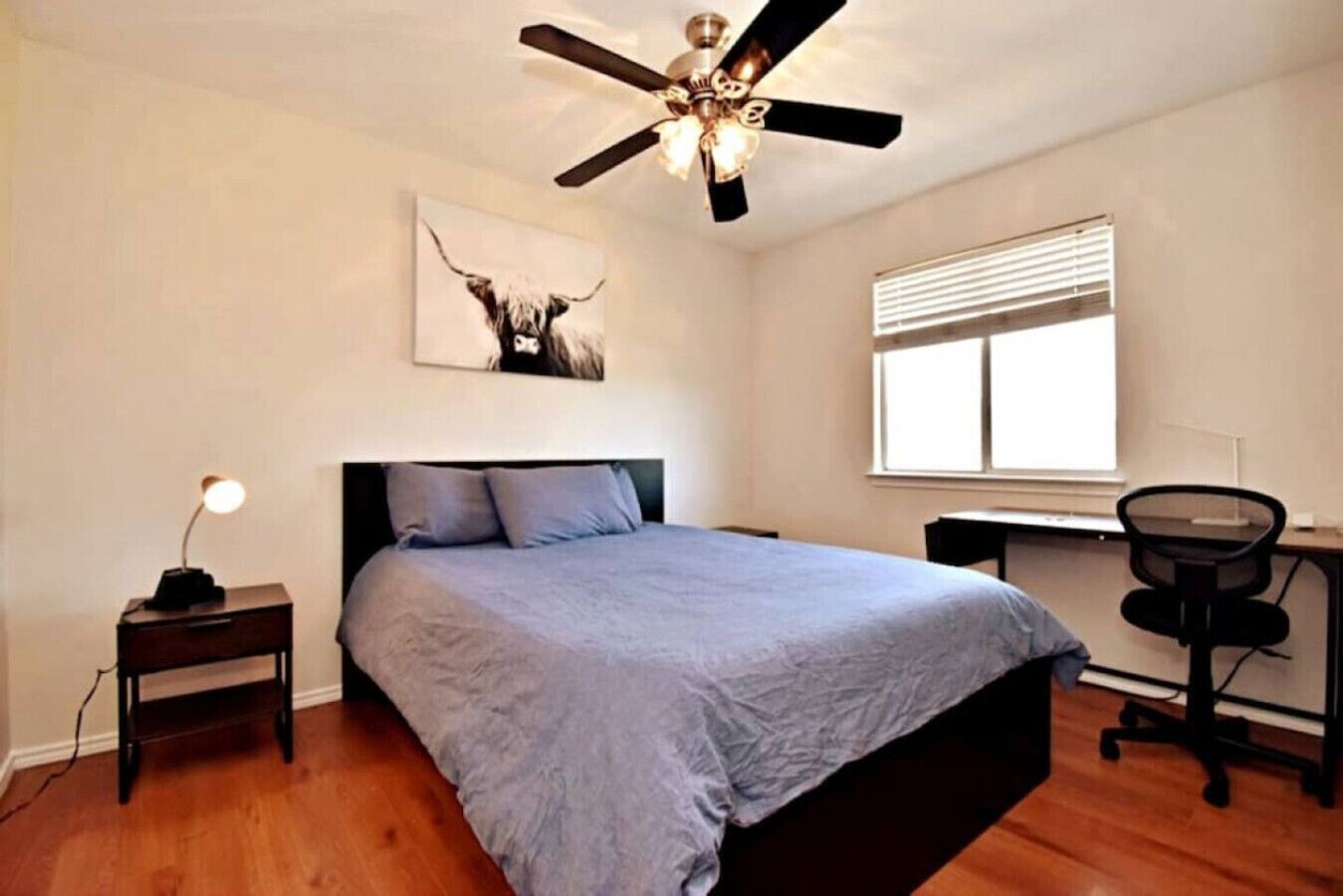Une chambre à coucher comprenant un lit, un bureau, une chaise, un ventilateur de plafond, une fenêtre avec des stores et une œuvre d’art accrochée au mur.