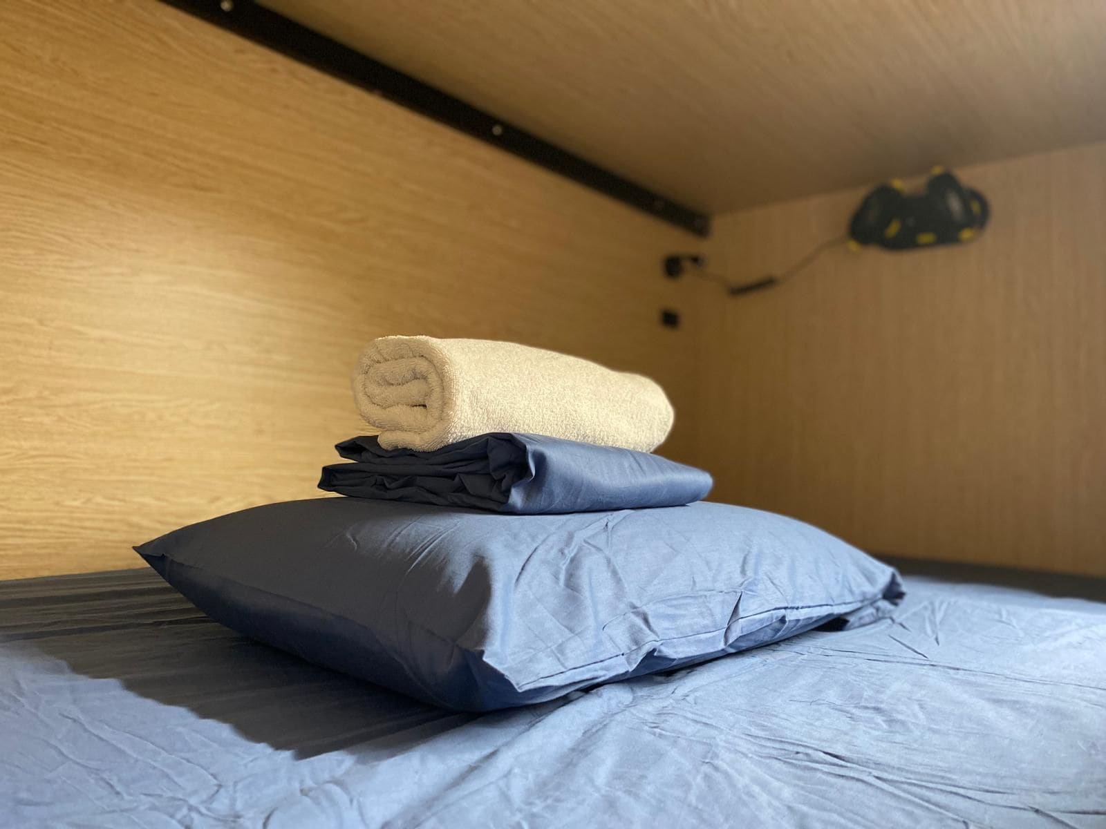 A bed with a rolled towel and pillows, a wooden wall, and a black object on the wall.