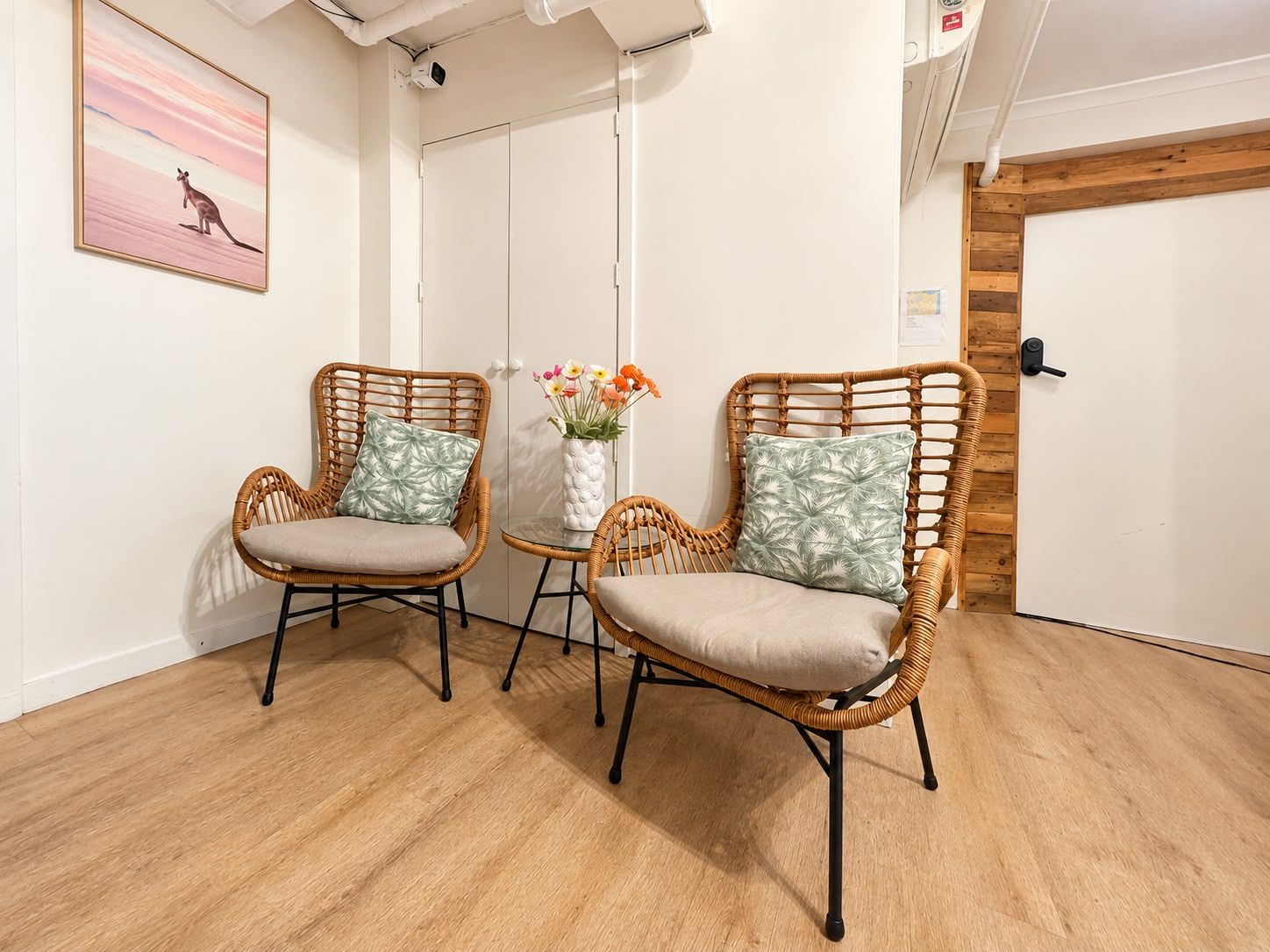 A room with two wicker chairs, a small table with a vase of flowers, and a painting on the wall.