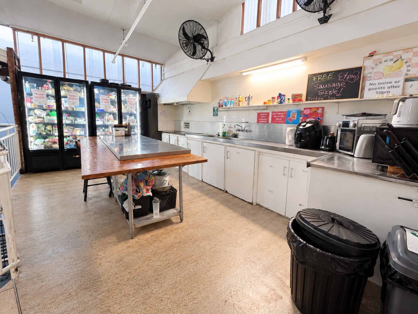 Una cocina con una mesa de madera, un fregadero y un refrigerador. Hay dos botes de basura y un ventilador en el techo.
