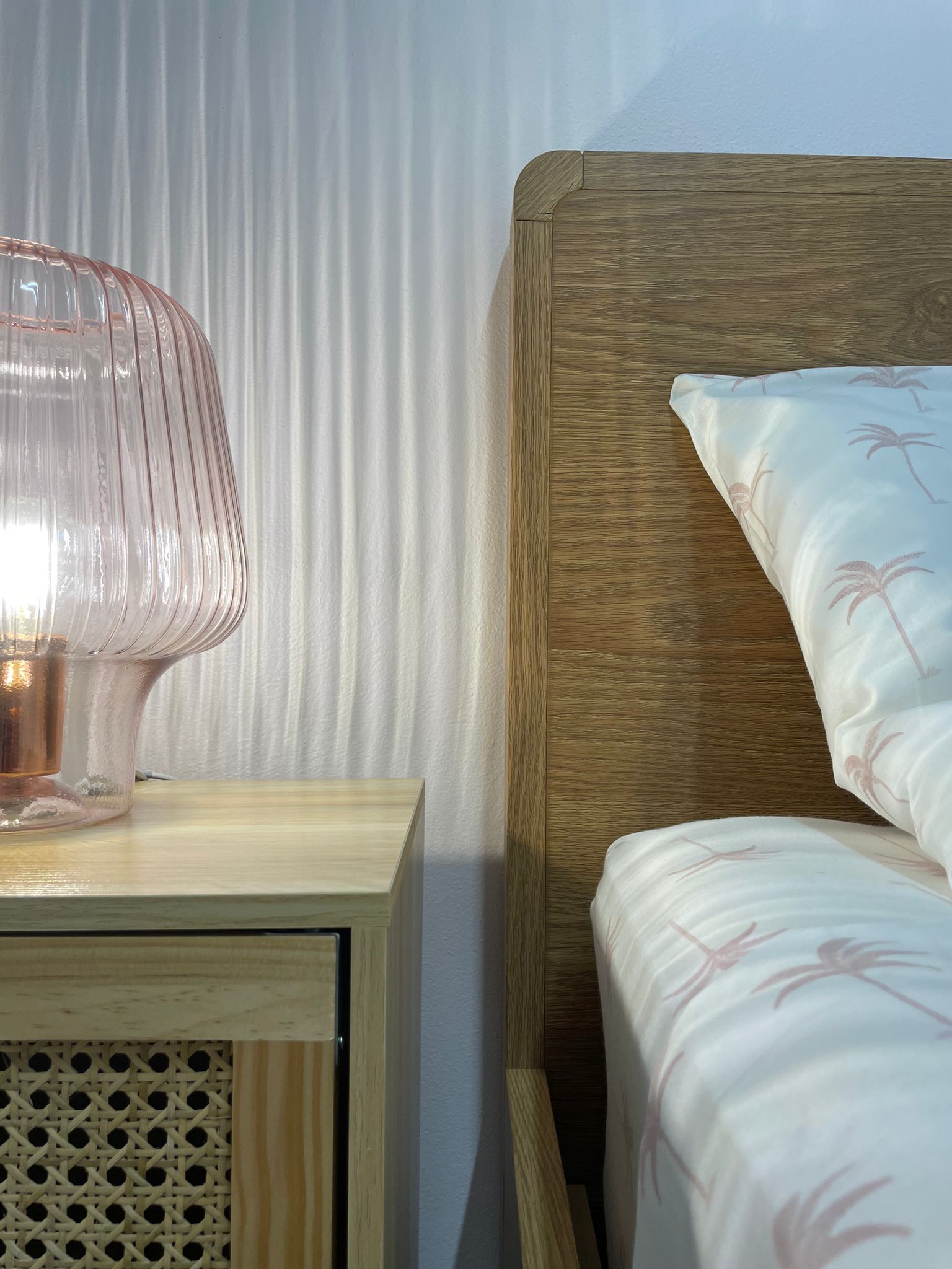 Cama com cabeceira de madeira e roupa de cama branca com estampas de palmeiras, mesa de cabeceira com abajur rosa e criado-mudo de madeira com painel frontal trançado.