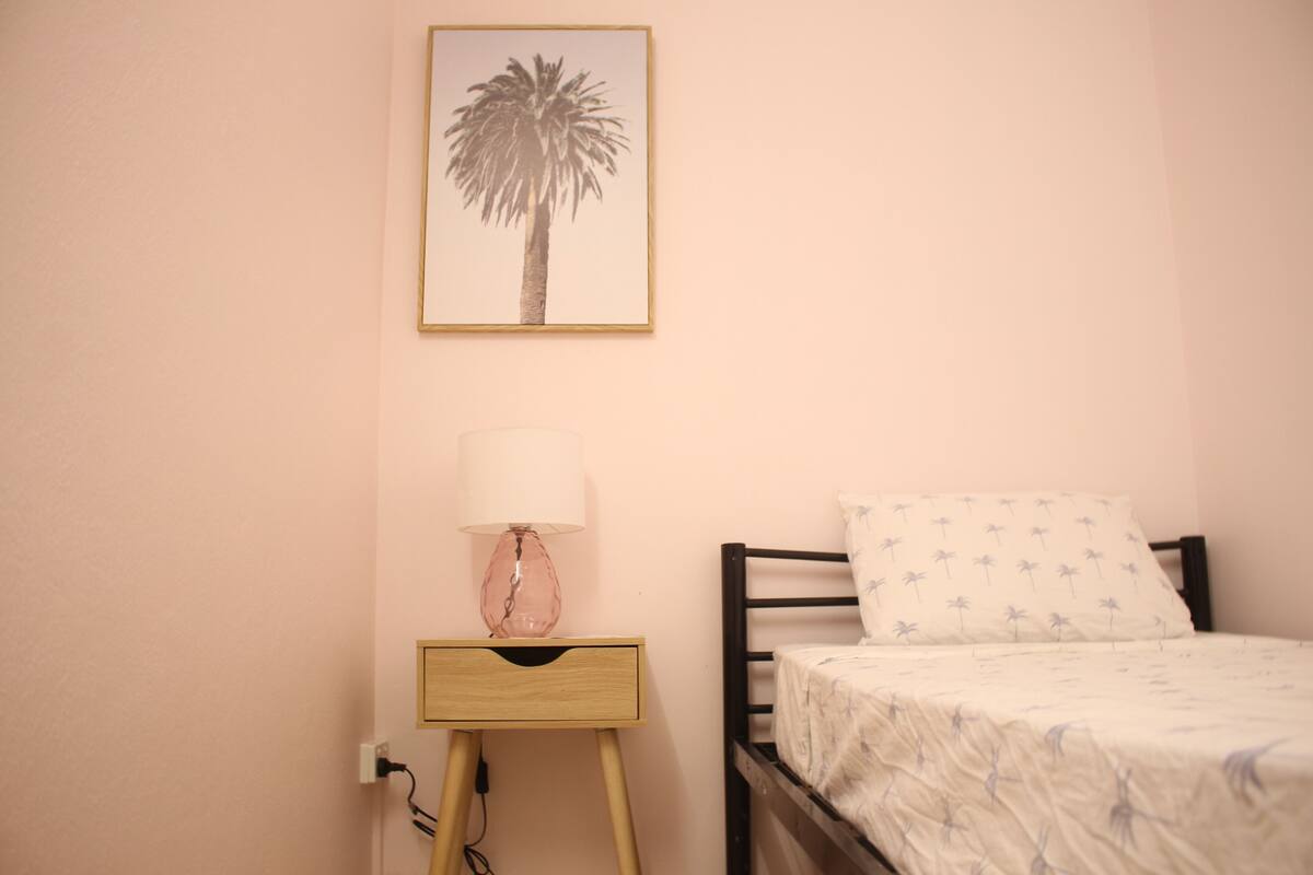 Un dormitorio con una cama, una mesita de noche con lámpara, un cuadro en la pared y un enchufe.