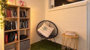 A room with a white brick wall, a bookshelf filled with books, a black wicker chair with a cushion, a small round table with a book, and a window with a view.