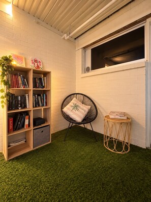 A room with a white brick wall, a bookshelf filled with books, a black wicker chair with a cushion, a small round table with a book, and a window with a view. - The Espy Hostel (Cairns)