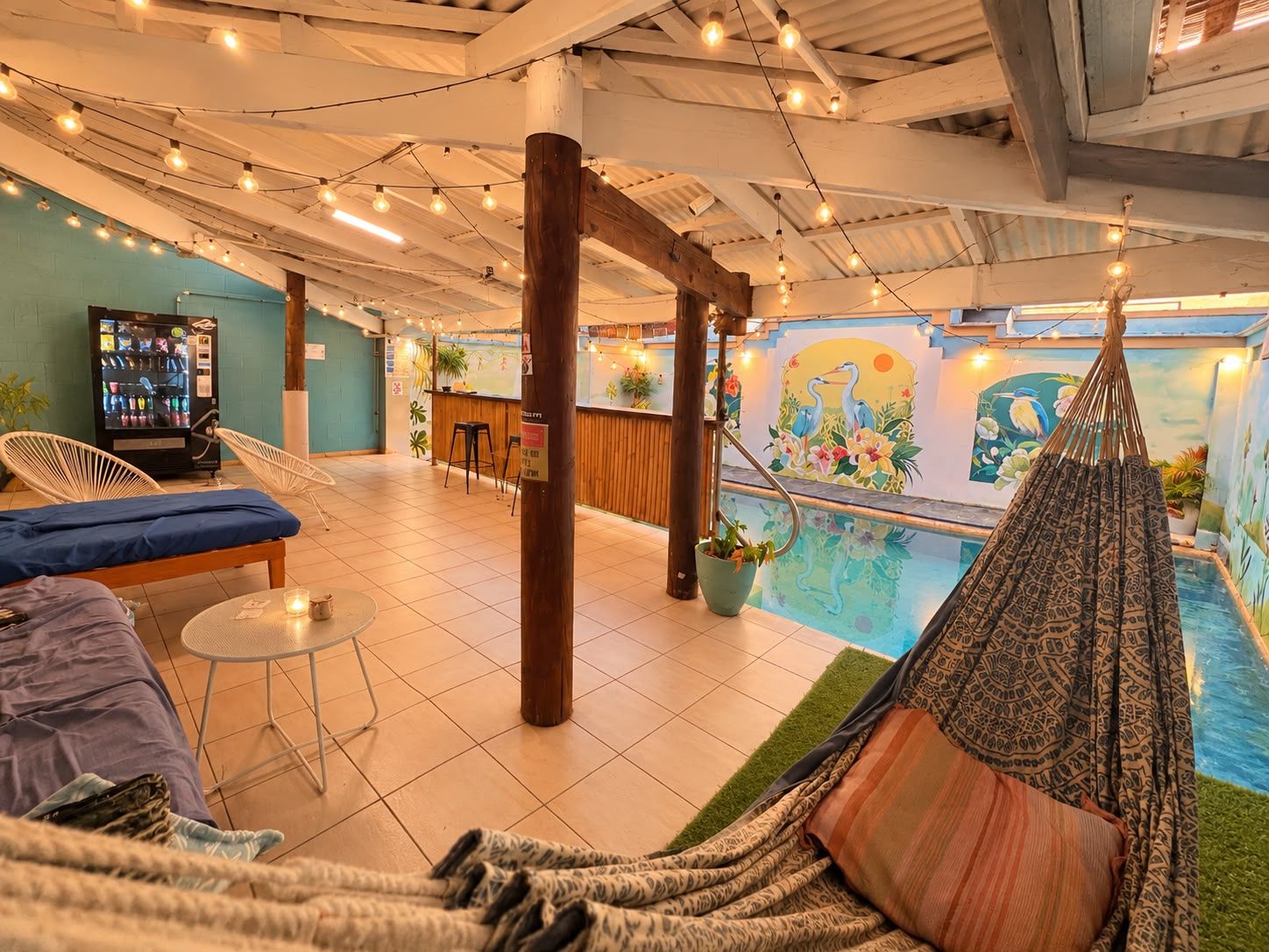 An indoor pool area with a hammock, lounge chairs, a bar, and a mural on the wall.