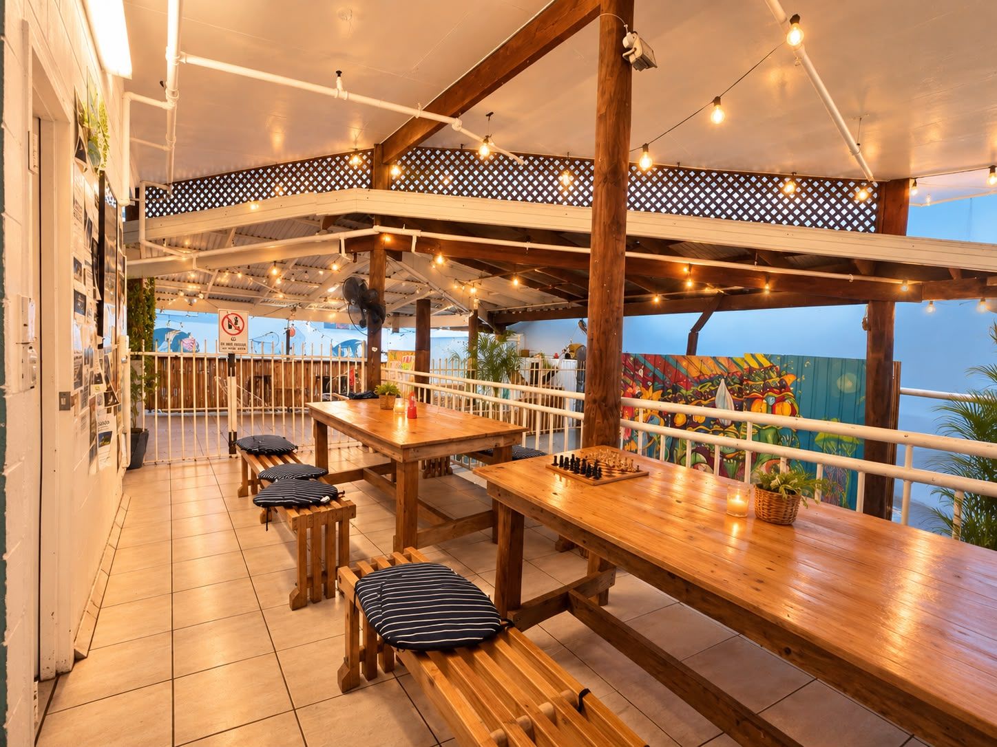 An outdoor seating area with wooden tables and benches, string lights, and a view of the sea.