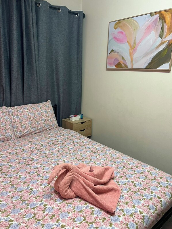 A bedroom with a floral bedspread, a pink towel, a nightstand with toiletries, and a painting on the wall. - The Espy Hostel (Cairns)