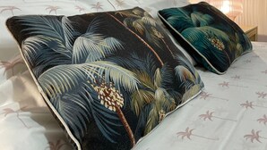 A bed with two decorative pillows featuring tropical leaf patterns, a wooden headboard, and a bedside lamp. - The Espy Hostel (Cairns)
