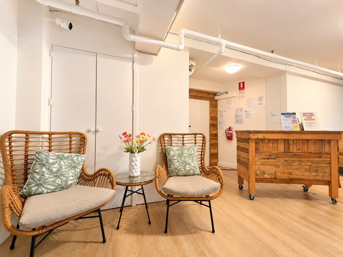 A room with two wicker chairs, a small table with a vase of flowers, and a wooden desk with a sign.