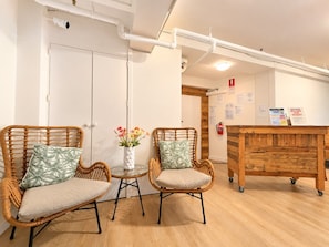 A room with two wicker chairs, a small table with a vase of flowers, and a wooden desk with a sign. - The Espy Hostel (Cairns)