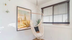A room with a hammock chair, a large framed painting, and a window with blinds. - ✧VIEWS✧ 6min to Joshua Tree walk to bars + firepit  (Twentynine Palms)