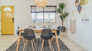 A dining area with a wooden table, black chairs, a patterned rug, a yellow door, and a large window with a view. - ✧VIEWS✧ 6min to Joshua Tree walk to bars + firepit  (Twentynine Palms)